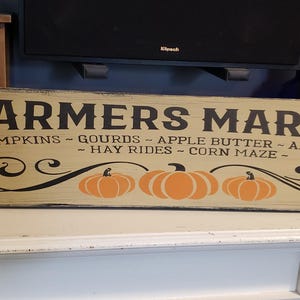 Könnte beinhalten: Ein rustikales Holzschild mit dem Text "FARMERS MARKET" in schwarzen Buchstaben. Das Schild ist in einem hellen Beige gestrichen und hat ein dekoratives Rankendesign mit drei orangefarbenen Kürbissen. Der Text unter dem Schild lautet "PUMPKINS - GOURDS - APPLE BUTTER - APPLE CIDER - HAY RIDES - CORN MAZE - "