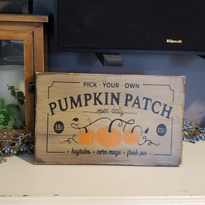 Op de afbeelding: Een rustiek houten bord met de tekst "Pick Your Own Pumpkin Patch - open daily - hayrides - corn maze - pumpkin pie" en een tekening van drie pompoenen aan een wijnstok.