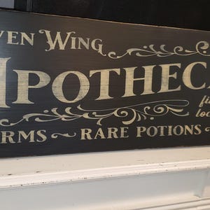 Op de afbeelding: Een zwart houten bord met witte letters waarop "Raven Wing Apothecary" staat, met daaronder de woorden "finest quality locally brewed" en "charms - rare potions - spells".