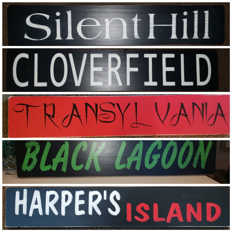 Op de afbeelding: Vijf houten borden met een zwarte achtergrond en witte letters. De borden lezen "Silent Hill", "Cloverfield", "Transylvania", "Black Lagoon" en "Harper's Island".