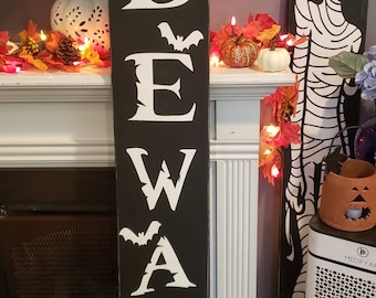 May include: A black wooden sign with white letters spelling "BEWARE" with a bat silhouette between each letter. The sign is leaning against a white fireplace mantel.