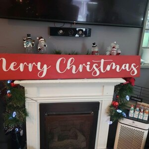 May include: A red wooden sign with the words "Merry Christmas" in white cursive lettering. The sign is sitting on a white mantel above a fireplace.
