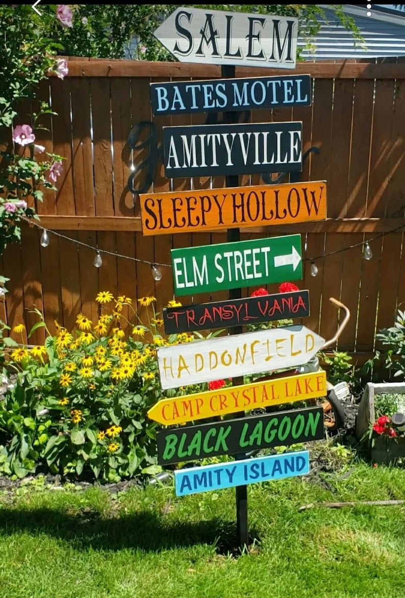 Op de afbeelding: Een houten wegwijzer met meerdere richtingborden die naar verschillende fictieve locaties uit horrorfilms wijzen. De borden zijn in verschillende kleuren geschilderd en dragen de namen: Salem, Bates Motel, Amityville, Sleepy Hollow, Elm Street, Transsylvani&euml;, Haddonfield, Camp Crystal Lake, Black Lagoon en Amity Island.