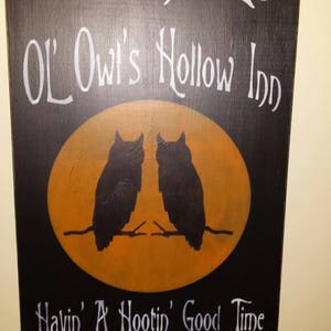 Puede incluir: Un letrero de madera negro con texto blanco que dice "Welcome to Ol' Owl's Hollow Inn Havin' A Hootin' Good Time Since 1787". El letrero presenta un círculo amarillo con dos siluetas de búhos negros.
