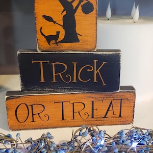 Puede incluir: Bloques de madera con temática de Halloween que deletrean "TRICK OR TREAT". El bloque superior presenta una silueta de bruja con un gato. Los bloques son de color naranja y negro con un acabado envejecido. Una guirnalda de bayas azules con luces está en primer plano.