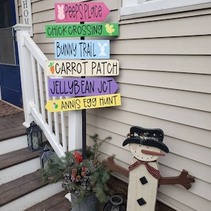 Op de afbeelding: Een houten wegwijzer met zes kleurrijke pijlen die in verschillende richtingen wijzen. Elke pijl heeft een ander paasbericht, waaronder "Peeps Place", "Chick Crossing", "Bunny Trail", "Carrot Patch", "Jellybean Jct" en "Annis Egg Hunt".