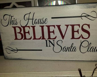 May include: White wooden sign with black and red lettering that reads "This House Believes in Santa Claus". The sign has a distressed look and is decorated with black swirls.
