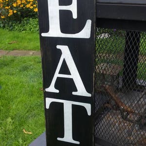 May include: A black wooden sign displaying the word "EAT" in large, white capital letters. The sign has a rectangular shape and a distressed finish. The background features green grass and yellow flowers.