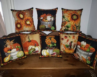 Può includere: Otto cuscini decorativi con motivi autunnali. I cuscini presentano zucche, girasoli, zucche e foglie in tonalità di arancione, giallo, verde e marrone. I cuscini sono disposti su una superficie di legno.