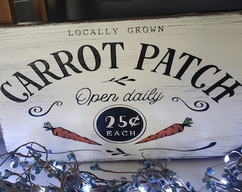 May include: White wooden sign with black text that reads "Locally Grown Carrot Patch Open Daily 25¢ Each". The sign has a distressed look and features two orange carrots with green tops.