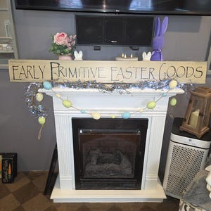 Puede incluir: Chimenea blanca con interior negro, decorada para Semana Santa. Un letrero de madera dice "EARLY PRIMITIVE EASTER GOODS". La repisa está adornada con una guirnalda de huevos de Pascua y un conejito morado. También hay un arreglo floral rosa y velas.