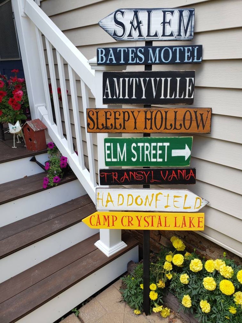 Op de afbeelding: Een houten wegwijzer met meerdere richtingspijlen die naar verschillende locaties uit horrorfilms wijzen. De borden geven aan: Salem, Bates Motel, Amityville, Sleepy Hollow, Elm Street, Transsylvani&euml;, Haddonfield en Camp Crystal Lake.