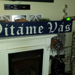 May include: A blue wooden sign with white lettering that reads "Vitáme Vás". The sign is hanging above a white fireplace.
