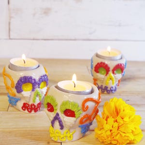 Può includere: Tre portacandele a forma di teschio in ceramica bianca con disegni floreali colorati e candele accese. I teschi hanno accenti rossi, verdi, viola e gialli. Un fiore giallo brillante è accanto ai portacandele.