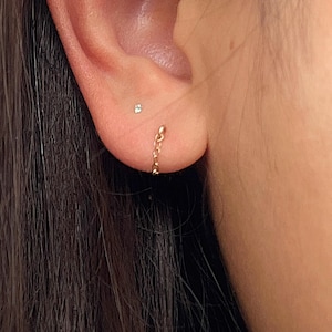 May include: A close-up of a person's ear adorned with two delicate gold earrings. One is a small stud, and the other is a chain earring that dangles gracefully. The earrings add a touch of elegance to the ear.