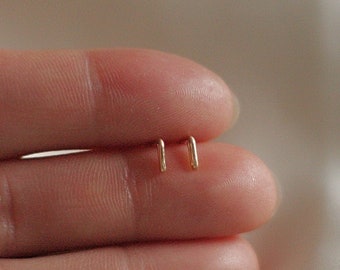 May include: A pair of small, gold-colored, minimalist earrings. Each earring has a simple, straight bar shape with a slight curve at the end. The earrings are displayed on a person's finger against a soft, white background.