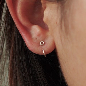 Puede incluir: Primer plano de una oreja con dos pendientes pequeños de plata. Uno es un pequeño botón y el otro un pendiente de aro fino. La piel es clara y el fondo es de un color suave y neutro.