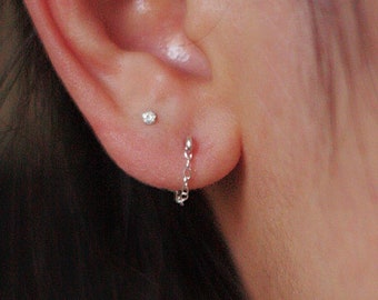 May include: A close-up of an ear with two earrings. One is a small stud, and the other is a delicate chain earring. The chain earring hangs down from the earlobe. The skin is light-toned.