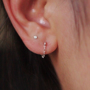 May include: A close-up of an ear with two earrings. One is a small stud, and the other is a delicate chain earring. The chain earring hangs down from the earlobe. The skin is light-toned.