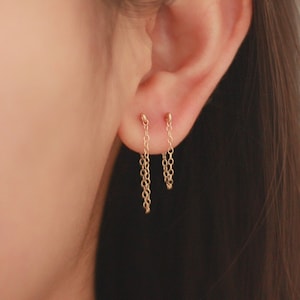 Puede incluir: Un par de delicados pendientes de cadena dorada. Un pendiente presenta una cadena más larga, mientras que el otro tiene una cadena más corta. Los pendientes se muestran en una oreja, con la cadena colgando hacia abajo.