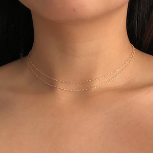 Puede incluir: Un collar de cadena de oro con dos cadenas delicadas superpuestas. El collar lo lleva una persona con el pecho descubierto y el pelo oscuro.