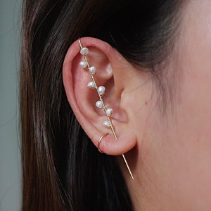 Puede incluir: Un pendiente de oreja dorado con un delicado dise&ntilde;o de peque&ntilde;as perlas blancas. El pendiente de oreja se curva a lo largo de la oreja, con una barra recta que se extiende hacia abajo. La joya se lleva en la oreja.