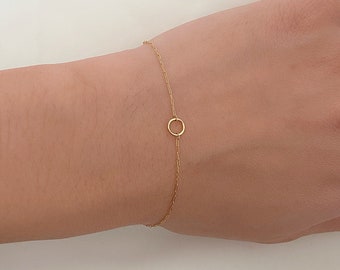 May include: Delicate gold bracelet featuring a small, open circle pendant. The bracelet is a fine chain, and the pendant is centered on the wrist. The jewelry is displayed on a light background.