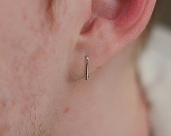 May include: A small, silver bar earring is worn in the ear. The earring is a simple, minimalist design, reflecting a modern aesthetic. The earring is a single, straight bar, showcasing a sleek and understated style. The earring is made of a shiny, metallic material.