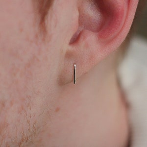 以下が含まれることがあります： 耳に装着された小さな銀色のバーピアス。ピアスはシンプルでミニマリストなデザインで、モダンな美学を反映しています。ピアスは一本のまっすぐなバーで、洗練された控えめなスタイルを表現しています。
