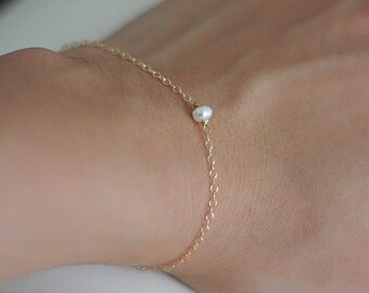 May include: Delicate gold chain bracelet featuring a single, small white pearl. The bracelet is simple and elegant, perfect for everyday wear. The chain is thin and the pearl is centered on the bracelet.