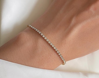 May include: A gold tennis bracelet with a row of sparkling, clear gemstones. The bracelet is worn on a wrist, against a backdrop of a white fabric. The bracelet appears to be delicate and elegant, suitable for formal occasions.