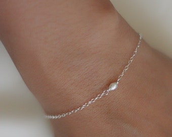 May include: A delicate silver bracelet featuring a small, oval-shaped pearl. The bracelet is a simple chain design, perfect for everyday wear. The pearl is centered on the chain, adding a touch of elegance to the minimalist style.