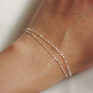 Könnte beinhalten: Zwei silberne Kettenarmbänder, die an einem Handgelenk übereinander liegen. Das obere Armband ist eine zarte Kette mit einer etwas größeren Gliedgröße. Das untere Armband ist eine dünnere, zartere Kette.