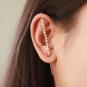Könnte beinhalten: Ein goldfarbener Ohrschmuck mit einer zarten Kette aus kleinen, weißen Perlen. Der Ohrring verläuft entlang des Ohrrandes, mit einem geraden Stab, der nach unten verläuft. Das Design ist schlicht und elegant.
