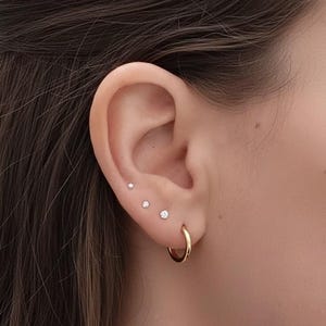 Può includere: Primo piano di un orecchio adornato da tre piccoli orecchini a bottone scintillanti e un orecchino a cerchio dorato. I borchie sono di dimensioni graduate. Il cerchio d'oro è un design semplice e classico.