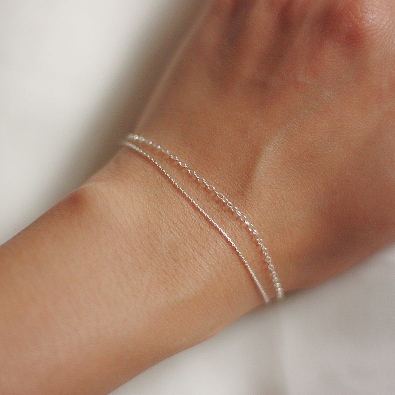 May include: Two silver chain bracelets layered on a wrist. The top bracelet is a delicate chain with a slightly larger link size. The bottom bracelet is a thinner, more delicate chain.
