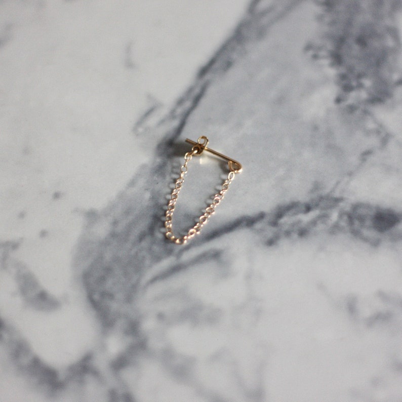 May include: A single gold-colored earring with a chain. The earring has a post for insertion into the ear and a delicate chain that dangles below. The earring is set against a marble background.
