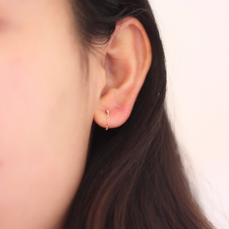 May include: A close-up of a gold chain earring. The earring features a small stud at the earlobe and a delicate chain that hangs down. The earring is a simple, elegant design.