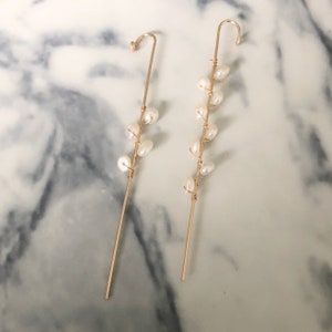 Puede incluir: Un par de trepadores de oreja dorados con un dise&ntilde;o delicado. Cada pendiente tiene un poste recto con una parte superior curvada y est&aacute; adornado con una serie de perlas blancas. Las perlas est&aacute;n unidas con alambre dorado, creando un aspecto elegante.