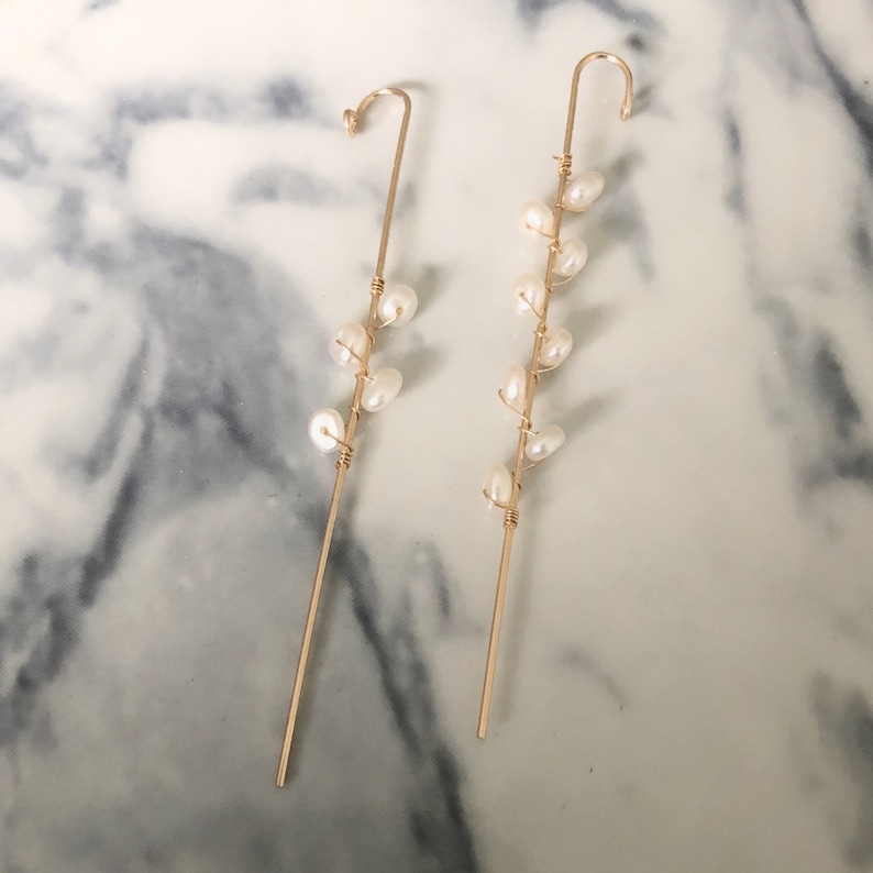 Puede incluir: Un par de trepadores de oreja dorados con un dise&ntilde;o delicado. Cada pendiente tiene un poste recto con una parte superior curvada y est&aacute; adornado con una serie de perlas blancas. Las perlas est&aacute;n unidas con alambre dorado, creando un aspecto elegante.