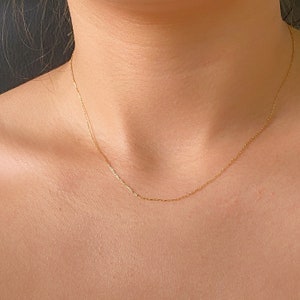 May include: A delicate gold necklace resting on a person's skin. The chain is thin and appears to be made of small, rectangular links. The necklace is centered and hangs just below the collarbone. The background is a dark, solid color.