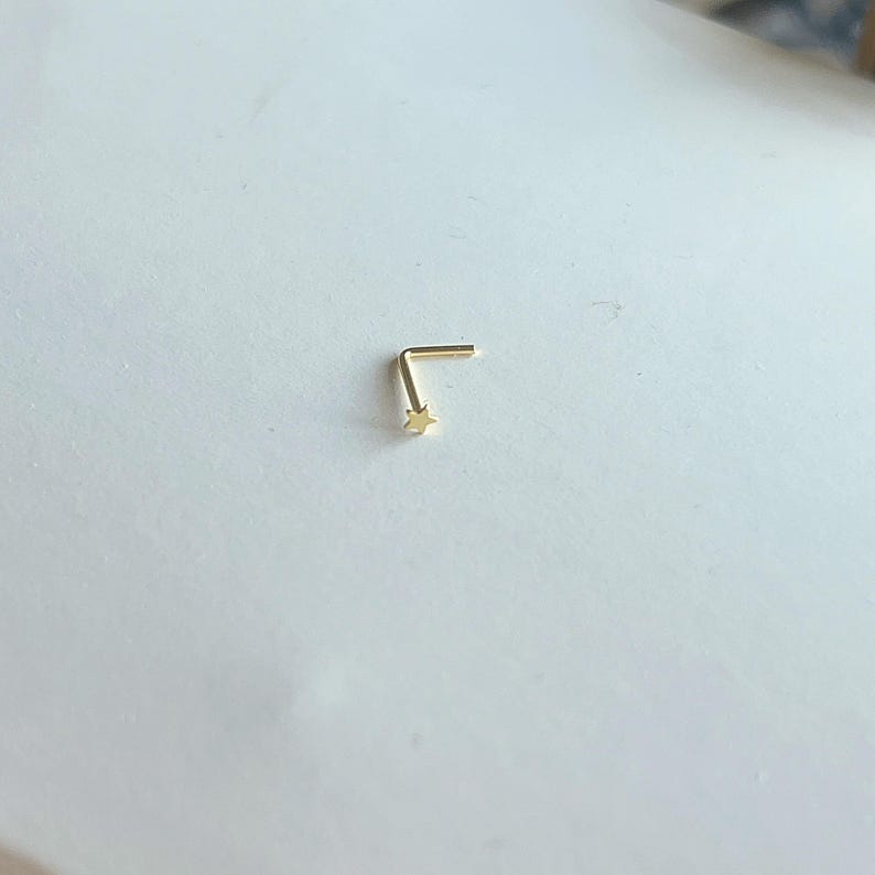 Puede incluir: Un peque&ntilde;o piercing de nariz dorado en forma de L. El pendiente presenta una peque&ntilde;a estrella en el extremo del lado m&aacute;s largo. La joya est&aacute; sobre un fondo blanco.