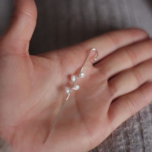 Puede incluir: Un pendiente de oreja plateado con un dise&ntilde;o delicado y peque&ntilde;os detalles de perlas blancas. El pendiente tiene un gancho curvado y un tallo delgado con tres perlas. La joya se sostiene en una mano.