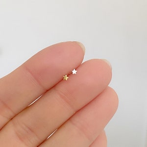 Puede incluir: Dos peque&ntilde;os pendientes en forma de estrella, uno dorado y otro plateado, se muestran en un dedo. Los pendientes tienen un dise&ntilde;o sencillo y delicado. El fondo es de un color suave y neutro.