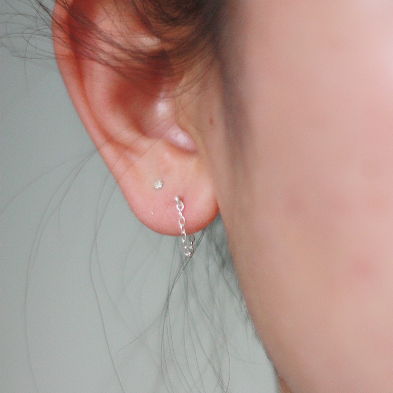 May include: A close-up of an ear with a small, clear stud earring and a silver chain earring. The chain earring dangles from the earlobe. The skin is light-toned.