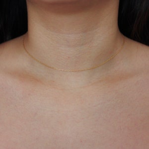 May include: A delicate gold chain necklace resting on a person's chest. The necklace is a simple, thin chain, likely made of gold, and sits close to the collarbone. The chain is a warm, yellow color.