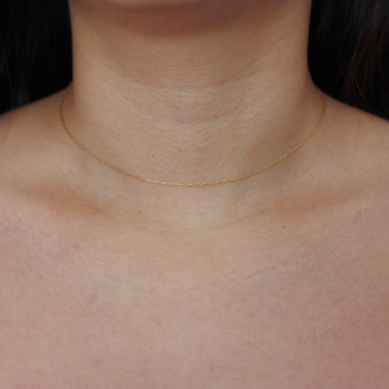 May include: A delicate gold chain necklace resting on a person's chest. The necklace is a simple, thin chain, likely made of gold, and sits close to the collarbone. The chain is a warm, yellow color.