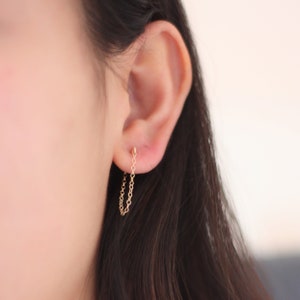 May include: A pair of delicate gold chain earrings. The earrings feature a small stud that connects to a dangling chain. The chain is made of small, interconnected links. The earrings are shown on an ear.