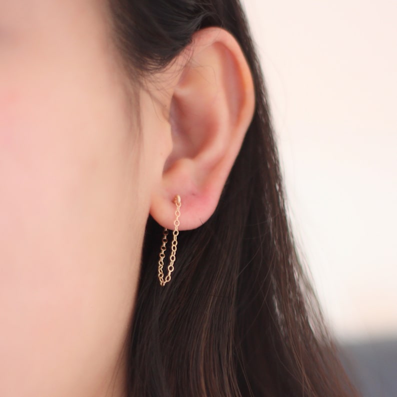 May include: A pair of delicate gold chain earrings. The earrings feature a small stud that connects to a dangling chain. The chain is made of small, interconnected links. The earrings are shown on an ear.