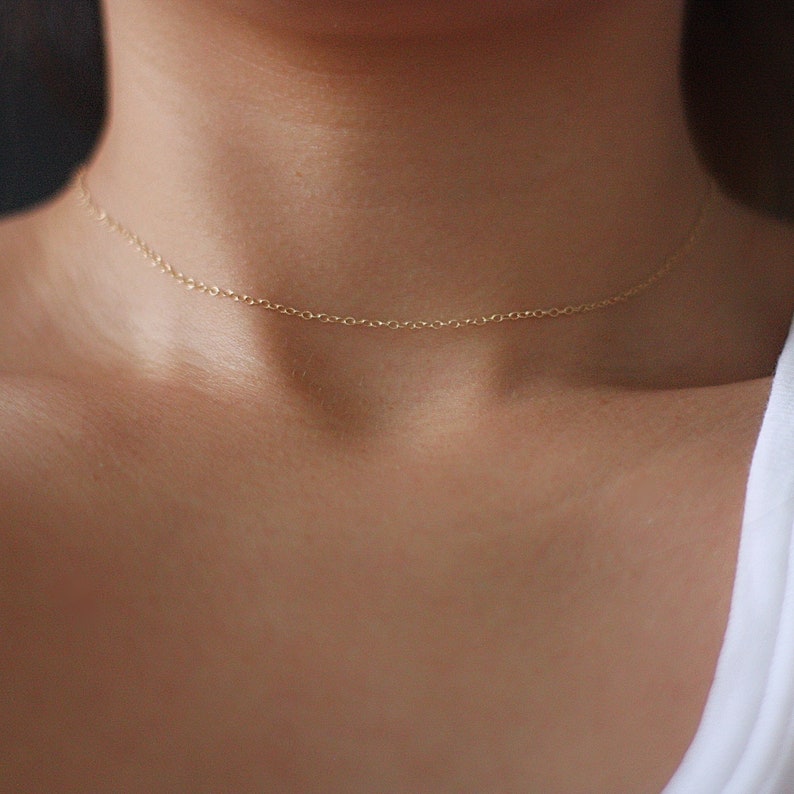 May include: A delicate gold chain necklace, worn as a choker, on a person's neck.
